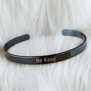 May include: A black metal bangle bracelet with the words "Be Kind" engraved on the front.