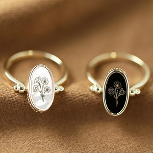 Puede incluir: Dos anillos de oro con engastes ovalados. Un anillo tiene un fondo blanco con un diseño floral negro, y el otro anillo tiene un fondo negro con un diseño floral dorado.