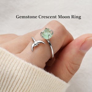 May include: A silver ring featuring a crescent moon design and a green gemstone. The ring is adjustable and can be worn on any finger.