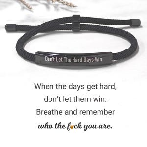 May include: Black bracelet with the inscription "Don't Let The Hard Days Win". The bracelet is crafted from a woven black cord and has a rectangular metal plate with engraved text. The image also includes motivational text.