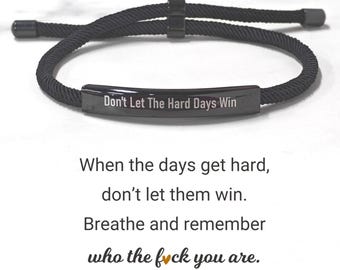 May include: Black bracelet with the text "Don't Let The Hard Days Win". The bracelet is made of a woven black cord and features a rectangular metal plate with engraved text. The image also includes inspirational text.
