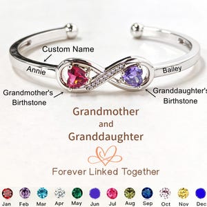 Könnte beinhalten: Ein silberfarbenes Armreif-Armband mit Unendlichkeitszeichen. Es hat einen herzförmigen roten Edelstein und einen herzförmigen lila Edelstein. Die Namen "Annie" und "Bailey" sind eingraviert. Der Text lautet "Großmutter und Enkelin für immer verbunden."