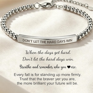 Puede incluir: Una pulsera plateada con pequeñas cuentas redondas y una barra central grabada con la frase "DON'T LET THE HARD DAYS WIN". La pulsera tiene una cadena y un cierre. Texto inspirador debajo.
