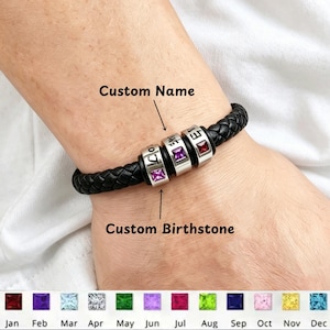 Op de afbeelding: Een zwarte gevlochten leren armband met drie zilverkleurige cilindrische kralen, elk ingelegd met een vierkante edelsteen. De kralen zijn gegraveerd met aangepaste namen en de tekst Custom Name en Custom Birthstone is zichtbaar.