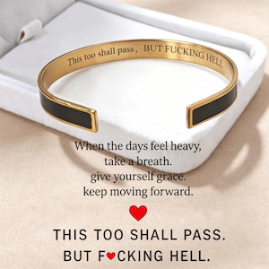 Könnte beinhalten: Ein goldfarbener Armreif mit schwarzer Emaille-Einlage. Die Innenseite ist mit dem Text graviert: This too shall pass, BUT FUCKING HELL. Der Armreif wird in einer weißen Schachtel präsentiert, mit zusätzlichem Text darunter über das Atmen.