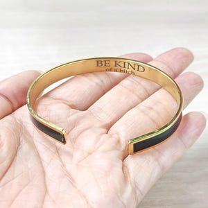 May include: Gold-tone cuff bracelet with black enamel accents. The bracelet features the engraved text "BE KIND ...of a bitch". The open-ended design allows for easy wear. A stylish accessory.