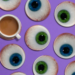 Könnte beinhalten: Eine weiße Teetasse mit Kaffee steht auf einer Untertasse, umgeben von augenförmigen Untersetzern mit blauen und grünen Iris, vor einem lila Hintergrund. Die Untersetzer haben ein realistisches Augendesign.
