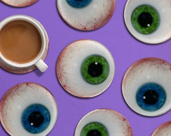 Puede incluir: Una taza blanca con café descansa sobre un platillo, rodeada de posavasos con forma de ojo con iris azules y verdes, sobre un fondo morado. Los posavasos tienen un diseño realista de ojo.