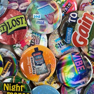 May include: A collection of colourful round buttons with various designs and text. Some of the buttons have a holographic finish. The text on the buttons includes "Get Lost!" "Your Pet" "More Coffee" "Nothing to Gain" "What Guilt?" "Be Happy!" "Night Moves" "Fragrance Your Fave" "Finish Quantum Jet-Dry" and "Vibe."