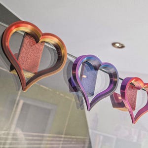 May include: Three heart-shaped decorations made of iridescent plastic. The hearts are layered on top of each other, with the largest heart at the back and the smallest heart at the front. The largest heart is red and gold, the middle heart is purple and blue, and the smallest heart is pink and orange.