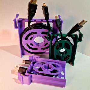以下が含まれることがあります： HDMIケーブル付きの紫と緑の3Dプリントケーブルオーガナイザー。オーガナイザーは、ケーブルを巻き付けるための溝のある円形デザインです。