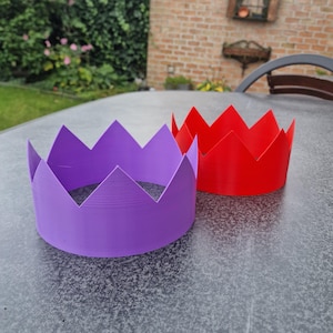 May include: Two colorful crowns, one purple and one red, sit on a gray surface. The crowns have a jagged, pointed design, resembling a royal crown. The background includes greenery and a brick wall.