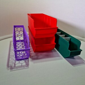 Peut inclure: Trois bacs de rangement en plastique, un violet, un rouge et un vert. Le bac rouge est empilé sur un autre bac rouge. Le bac vert a des séparateurs à l'intérieur.