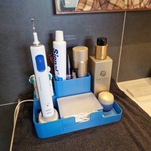 May include: A blue plastic bathroom organizer with compartments for toiletries. It contains a blue electric toothbrush, a tube of toothpaste, a bottle of lotion, a bottle of perfume, and a small container of cotton rounds. The organizer is sitting on a black towel.