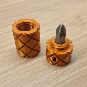 Puede incluir: Dos objetos marrones impresos en 3D con textura de piña. Uno es una funda cilíndrica, y el otro sostiene una punta de destornillador de metal oscuro. Los objetos están sobre una superficie de madera marrón claro.