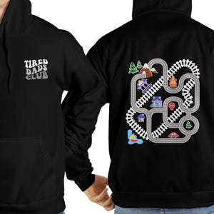 Pode incluir: Moletom preto com capuz com o texto "TIRED DADS CLUB" na frente. A parte de trás apresenta um gráfico colorido de uma pista de trem de brinquedo com casas, árvores e carros. O moletom tem um bolso frontal e um capuz com cordão.