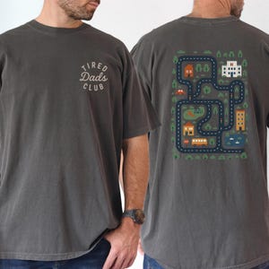May include: Dark grey t-shirt with the text "Tired Dads Club" on the front. The back of the shirt features a colourful graphic of a road map with houses, trees, and vehicles. The shirt is made of a soft, comfortable material.