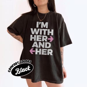 Op de afbeelding: Zwart Comfort Colors T-shirt met de witte tekst "I'M WITH HER AND HER" en roze pijlen. Het shirt heeft een relaxte pasvorm en een vintage look. De woorden "Comfort Colors" en "Black" staan in een zwart ovaal linksonder.
