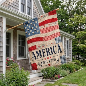 May include: A decorative American flag with a patchwork design, featuring blue stars, red stripes, and the word "AMERICA" with "EST. 1776". The flag is displayed outside a house, adding a touch of Americana.