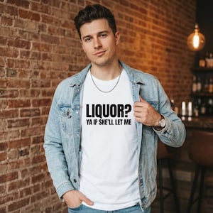 Op de afbeelding: Wit T-shirt met de zwarte tekst "LIQUOR? YA IF SHE'LL LET ME". De persoon draagt een lichtblauwe spijkerjas en een zilveren ketting. De achtergrond is een bakstenen muur.