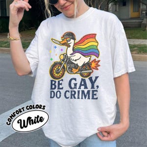 May include: White t-shirt with a graphic of a duck riding a motorcycle, wearing a helmet and rainbow cape, holding a knife. Text reads "BE GAY, DO CRIME." The shirt also has the words "COMFORT COLORS" and "White" printed on it.