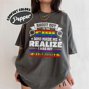 May include: A grey Comfort Colors t-shirt with the text "SHOUT OUT TO THE GIRL WHO MADE ME REALIZE I WAS NOT STRAIGHT" in white. The word "GIRL" is in rainbow colours. The shirt has a relaxed fit.