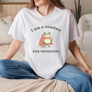 Peut inclure: T-shirt blanc avec le texte "I am a conduit FOR NONSENSE." Le t-shirt présente une illustration fantaisiste d'une grenouille portant une cape rouge et tenant une épée. La grenouille a de grands yeux expressifs.