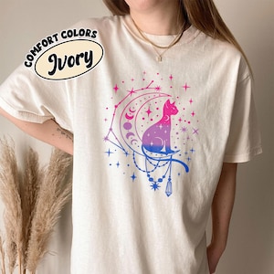 May include: Ivory t-shirt featuring a cat graphic on a crescent moon, in pink, purple, and blue hues. The shirt has the words "Comfort Colors" and "Ivory" printed on it.
