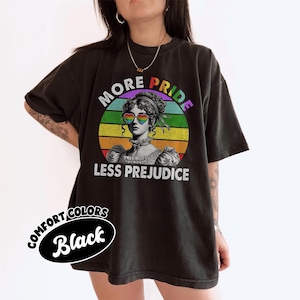 May include: Black t-shirt with a circular graphic of a woman wearing rainbow heart-shaped sunglasses, set against a rainbow background. The text reads "MORE PRIDE LESS PREJUDICE". The shirt also has the text "COMFORT COLORS Black".