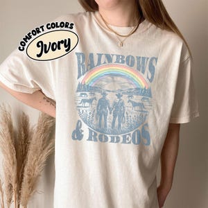 Peut inclure: T-shirt ivoire avec un motif représentant un arc-en-ciel, des cowboys, des chevaux et les mots "RAINBOWS & RODEO". Le t-shirt a un col rond et des manches courtes. Les mots "COMFORT COLORS" et "Ivory" sont imprimés en haut à gauche.