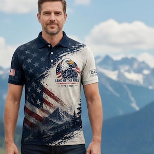 May include: A navy blue polo shirt with a patriotic design. The shirt features the American flag, an eagle, and the text "LAND OF THE FREE 1776-2026". The shirt has a white and blue color scheme and short sleeves.