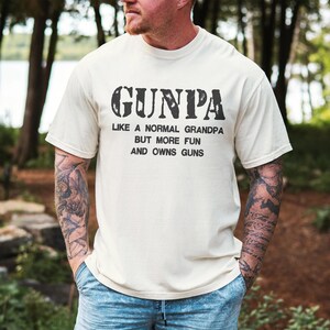 May include: Cream-colored t-shirt with the text "GUNPA" in a distressed font. Below, the shirt reads "LIKE A NORMAL GRANDPA BUT MORE FUN AND OWNS GUNS". The shirt is a casual style.