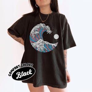 Op de afbeelding: Zwart Comfort Colors T-shirt met een gestileerd golfontwerp in blauw, roze en wit, met een witte maan en sterren. De tekst "Comfort Colors Black" staat linksonder.