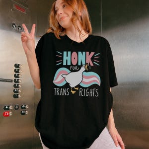 Può includere: T-shirt nero con la scritta "HONK FOR TRANS RIGHTS" e una grafica di un'oca bianca con dettagli rosa, blu e bianchi. La maglietta ha un girocollo e maniche corte. La persona nell'immagine indossa la maglietta.
