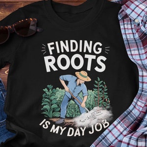 May include: Black t-shirt featuring a graphic of a person digging in a garden with the text "Finding Roots is My Day Job".