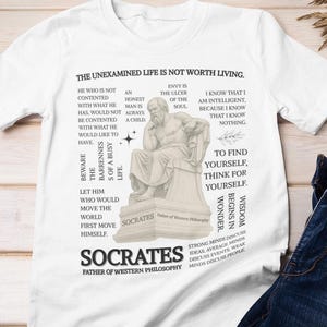 May include: White t-shirt featuring the quote "The Unexamined Life Is Not Worth Living" and a graphic of Socrates. The shirt includes additional text with philosophical quotes. A pair of blue jeans and a plaid shirt are also in the image.