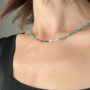 May include: A layered necklace featuring turquoise, pink, blue, and white beads, along with a delicate gold chain. The necklace is displayed on a person's neck, showcasing its colorful design and elegant style.