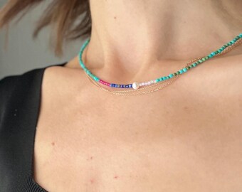 May include: A layered necklace featuring turquoise, pink, blue, and white beads, along with a delicate gold chain. The necklace is displayed on a person's neck, showcasing its colorful design and elegant style.