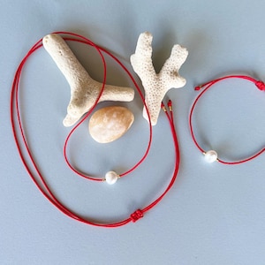 May include: A red cord necklace and bracelet set with a single white pearl. The jewelry is displayed with coral and a small, tan stone on a light blue background. The necklace is approximately 16 inches long.