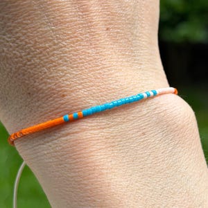 May include: A beaded bracelet with a colourful design. The bracelet features a pattern of orange, blue, and white beads. The bracelet is worn on a wrist, with a white string visible. The bracelet is a piece of jewellery.