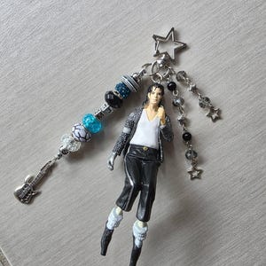 May include: A handmade keychain featuring a miniature figure of a pop star in a black and white outfit. The keychain includes a guitar charm, beads in shades of blue, black, and silver, and a star-shaped charm. The letters "MJ" are visible on the keychain.