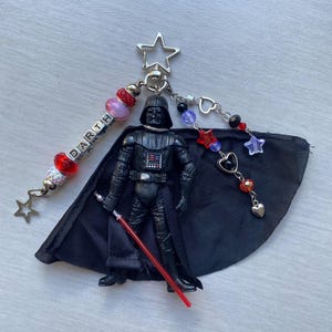 以下が含まれることがあります： 赤いライトセーバーを持った黒いダース・ベイダーのキーホルダー。キーホルダーには、フィギュア、黒いマント、そして「DARTH」と書かれたビーズチャームが含まれています。追加のチャームには、星、ハート、さまざまな色のビーズが含まれています。