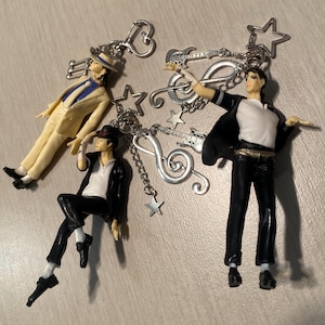May include: Four keychains featuring miniature figures of a performer in various poses. The figures are attached to silver-toned keychains with musical note and star charms. The figures are wearing black and white outfits.