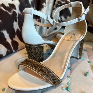 May include: White heeled sandals with a brown floral design on the straps and heel. The sandals have an ankle strap with a buckle closure. The brand name "Adalia" is printed on the insole.
