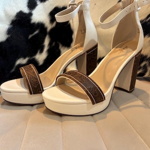 May include: A pair of platform heeled sandals. The sandals feature a cream-colored base and straps, with a brown, patterned leather accent on the heel and toe strap. The heels are approximately 4 inches high.