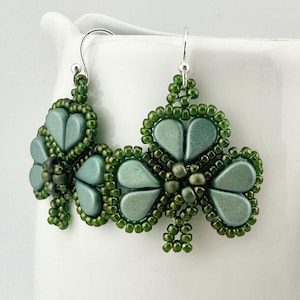 May include: A pair of handmade earrings featuring a shamrock design. The earrings are made with green beads and have a silver hook. The shamrock leaves are a muted teal color, and the edges are outlined with darker green beads.