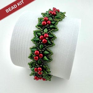 May include: A beaded bracelet with a holly berry design. The bracelet features dark green leaves and clusters of red beads. The words "BEAD KIT" are in a red banner in the upper left corner. The bracelet is displayed on a white surface.