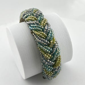 May include: Braided bracelet featuring a mix of silver, green, and gold seed beads. The bracelet is displayed on a white surface, showcasing its intricate woven design. The beads create a textured, eye-catching accessory.