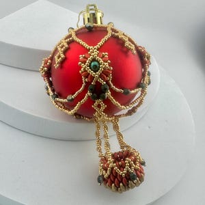 May include: A matte red spherical Christmas bauble decorated with intricate gold beadwork netting, featuring a central floral motif with a teal centre bead and a dangling beaded tassel element at the bottom, topped with a gold-coloured metal cap.