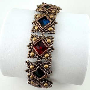 May include: A beaded bracelet with square, jewel-toned features in red, blue, and green. The bracelet has a bronze-coloured, intricate beadwork design with gold-coloured accents. The bracelet is displayed on a white surface.
